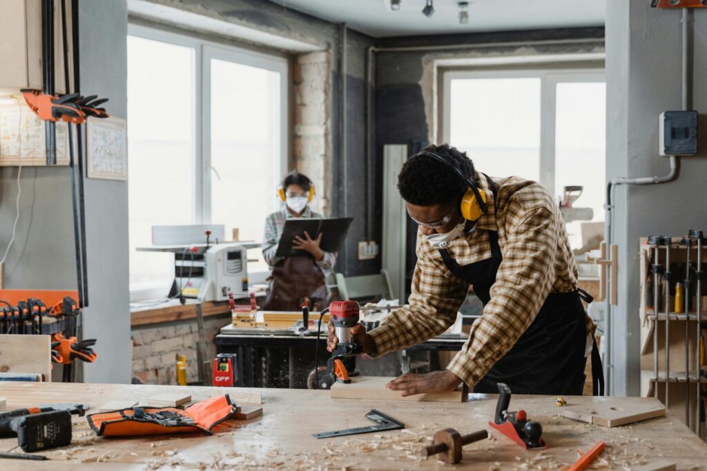 Carpenter using tools in a workshop with a colleague in the background.